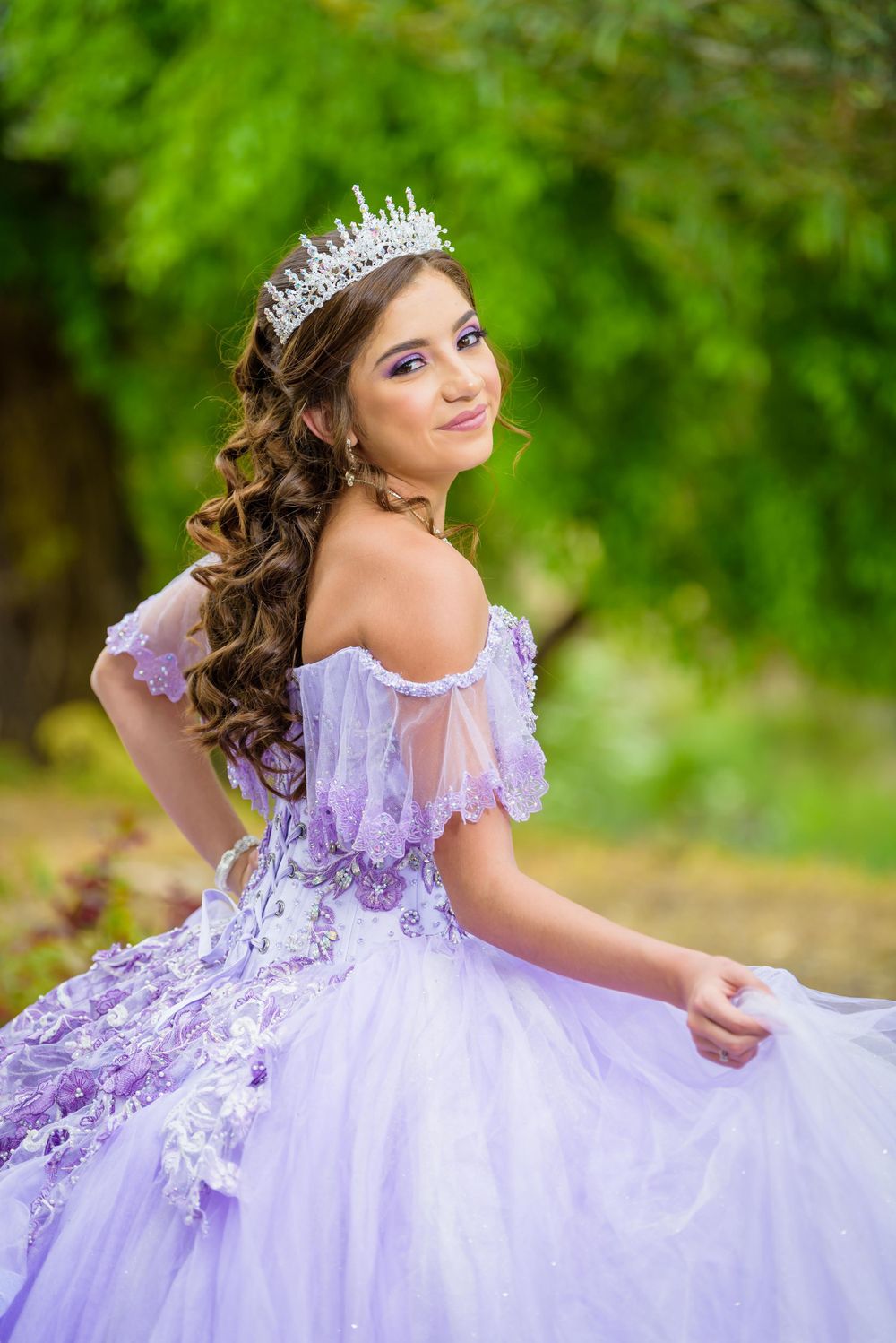 Young woman in lavender quinceañera ball gown with tiara poses outdoors against green foliage background.