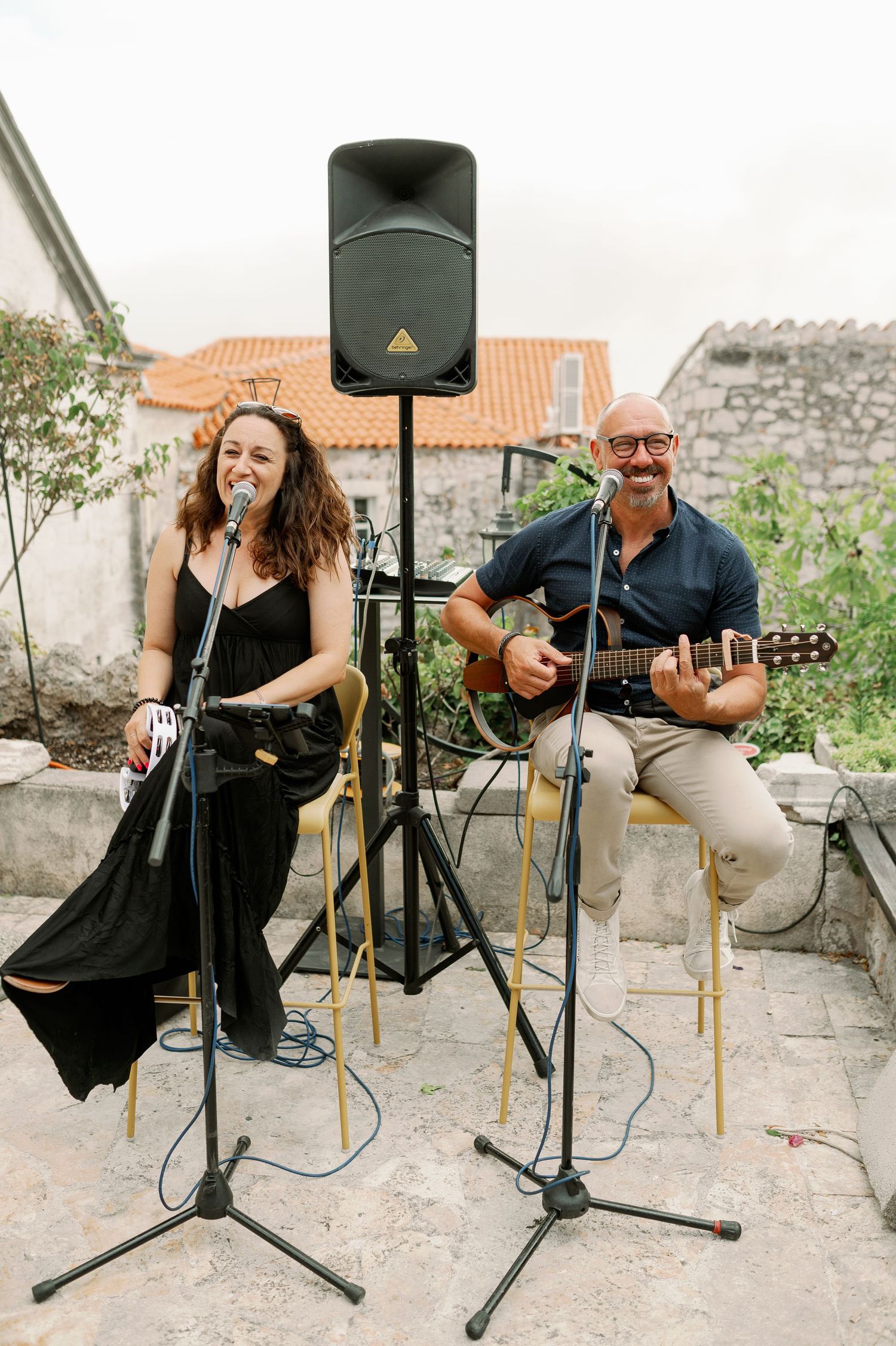 Musicians perform with acoustic guitar and microphone at an outdoor venue with stone walls and speaker setup.