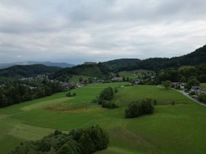 Panoramic aerial view of rolling green hills and mountains with scattered trees and a small village in the distance.