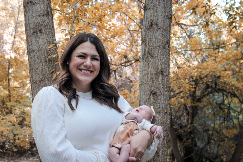 A person in white holds a baby while surrounded by fall foliage.