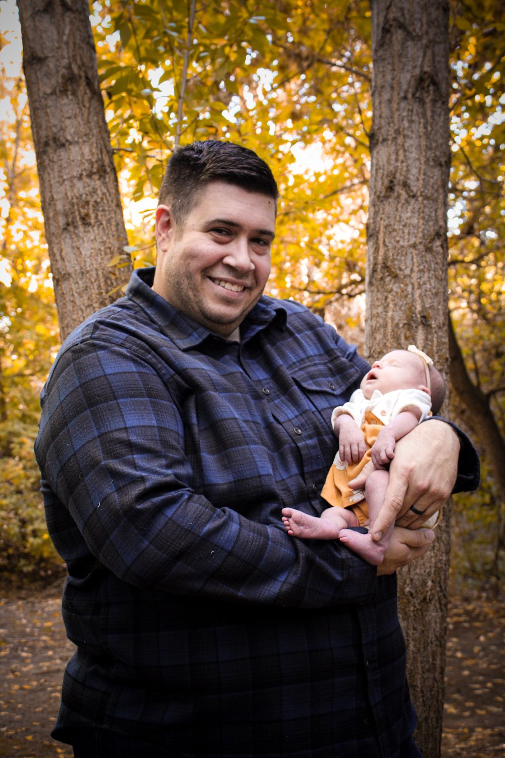 A person in a plaid shirt holds a small baby against a backdrop of autumn trees.