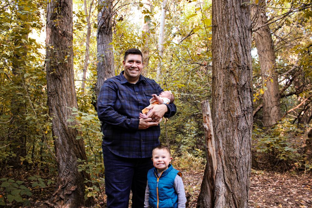 Three people stand between tall trees in a wooded area during autumn.