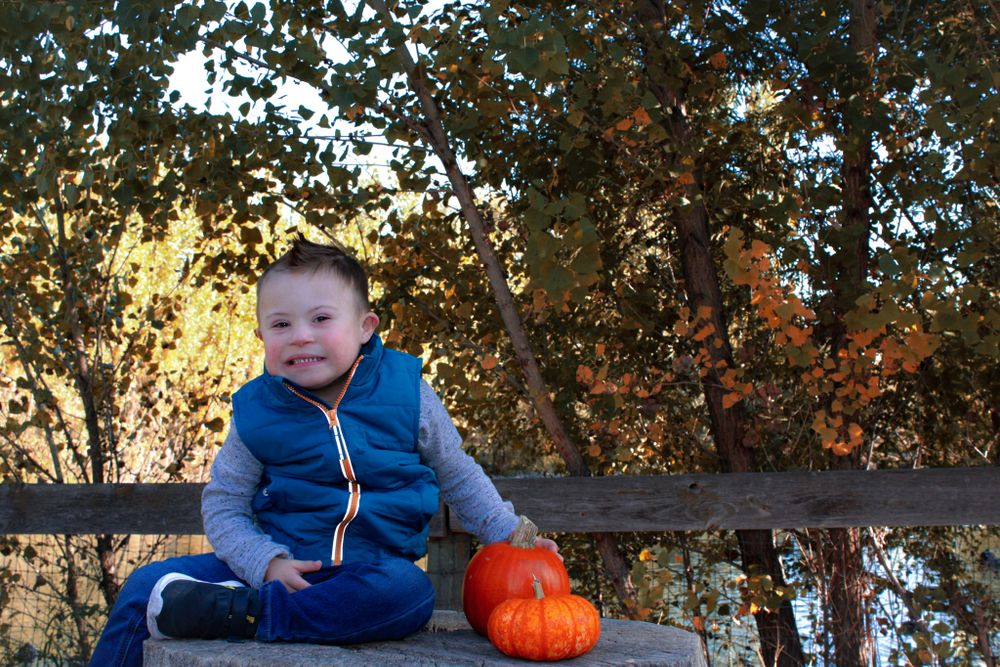 A toddler in blue sits next to an orange pumpkin on a wooden surface.