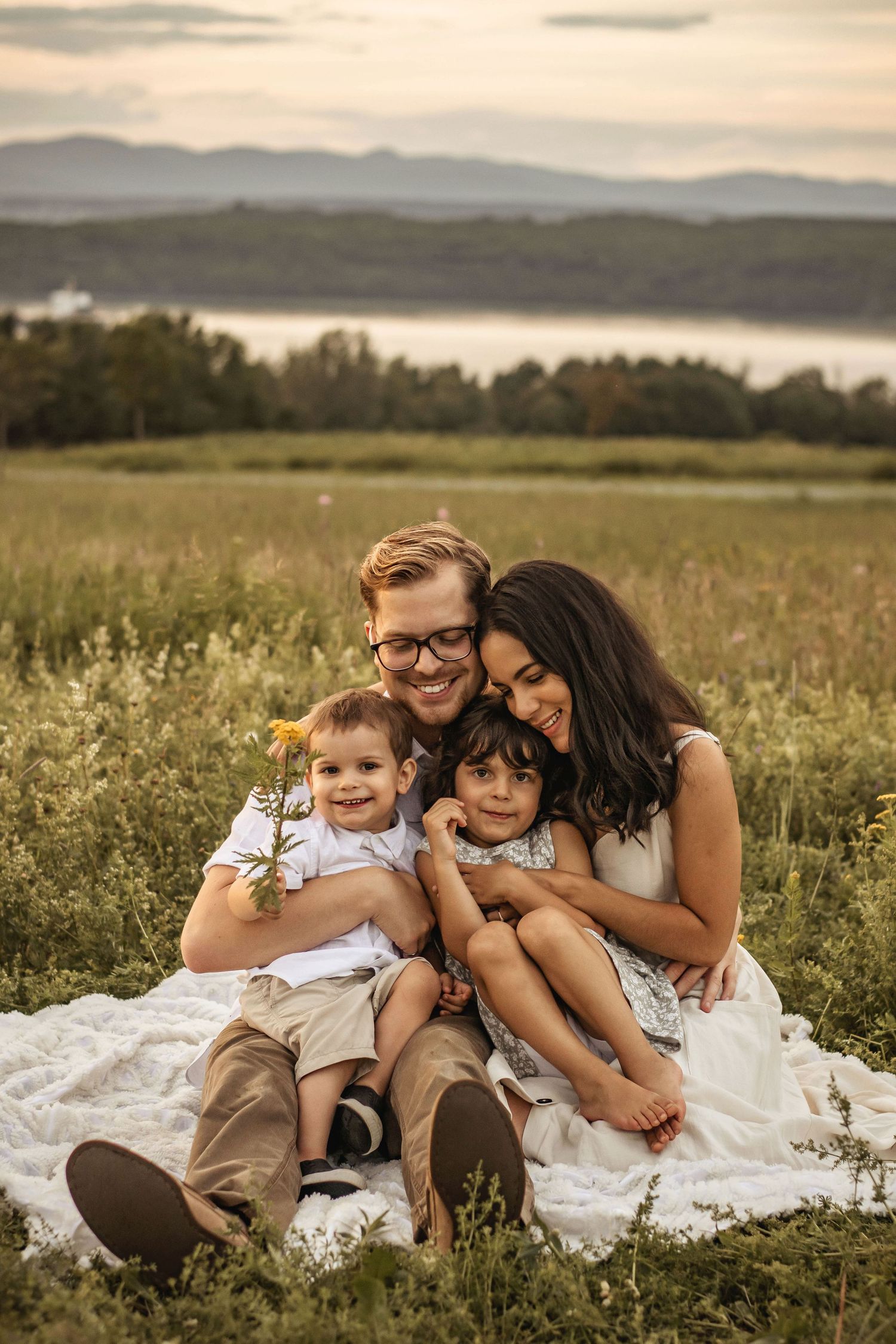 A family sits together on a blanket in a grassy field at sunset with mountains and a lake in the background.