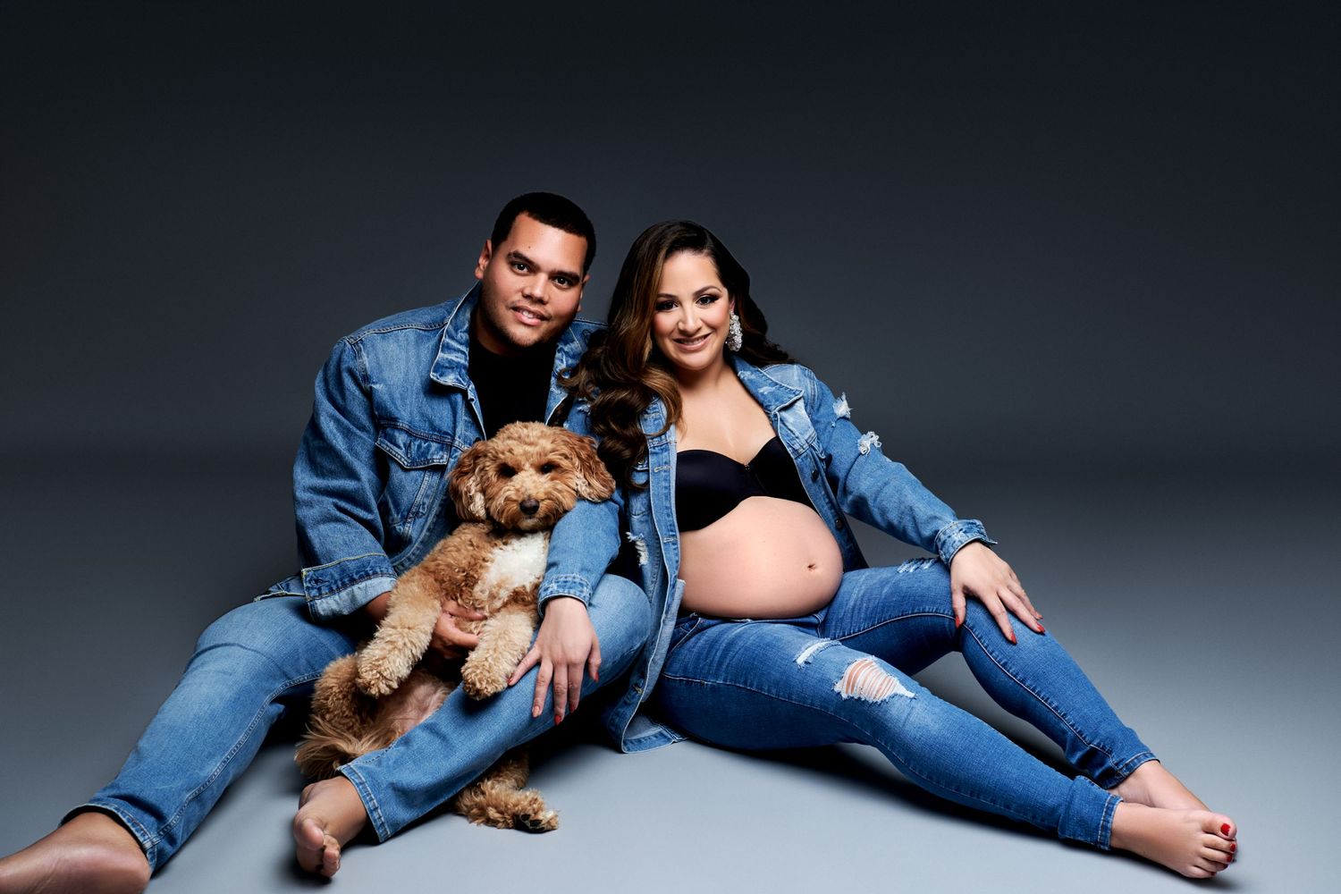 A pregnant woman and companion in matching denim outfits sit with a small brown dog against a dark gray backdrop. New Jersey Photographer