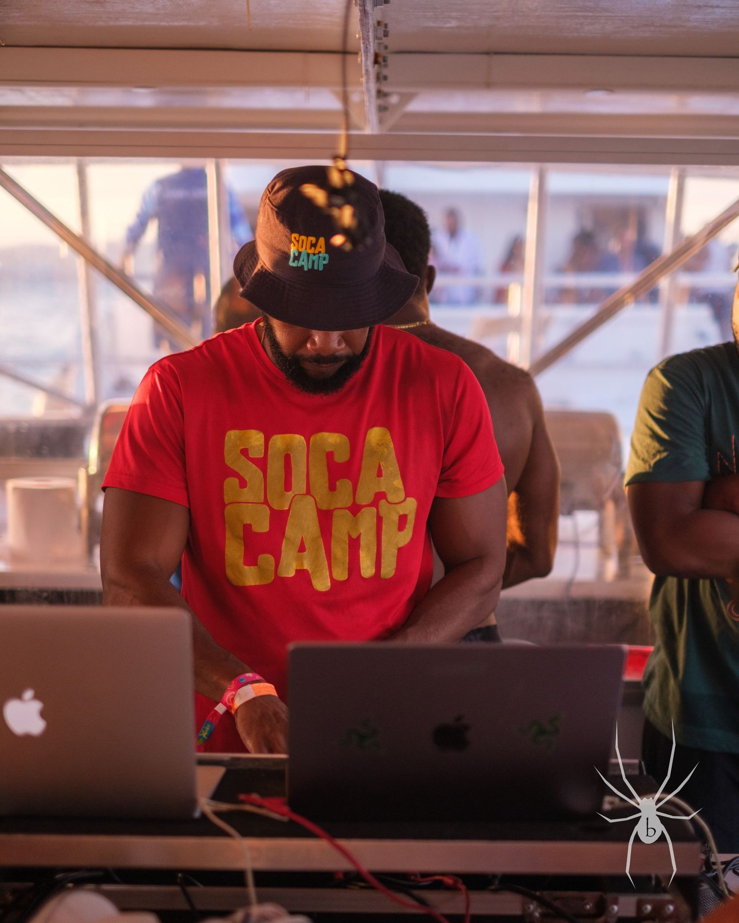 DJ wearing red SOCA CAMP t-shirt mixing music on laptops at an outdoor event.