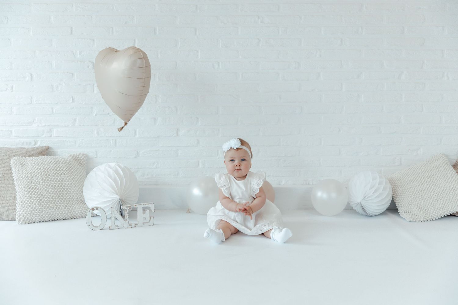 Baby in white outfit sitting among white balloons and heart decorations.