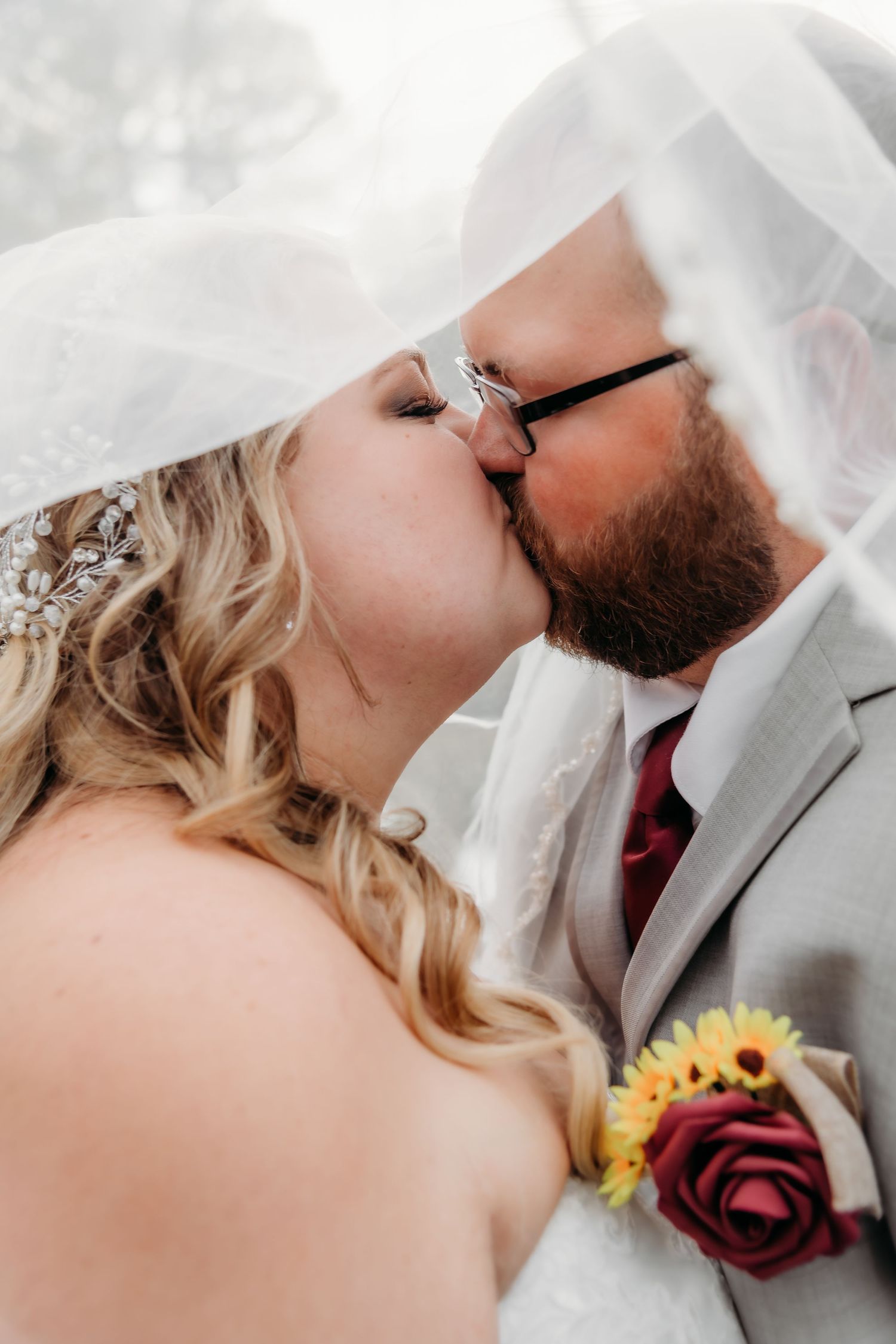 Intimate wedding kiss captured under flowing veil with sunflower and rose boutonniere detail.