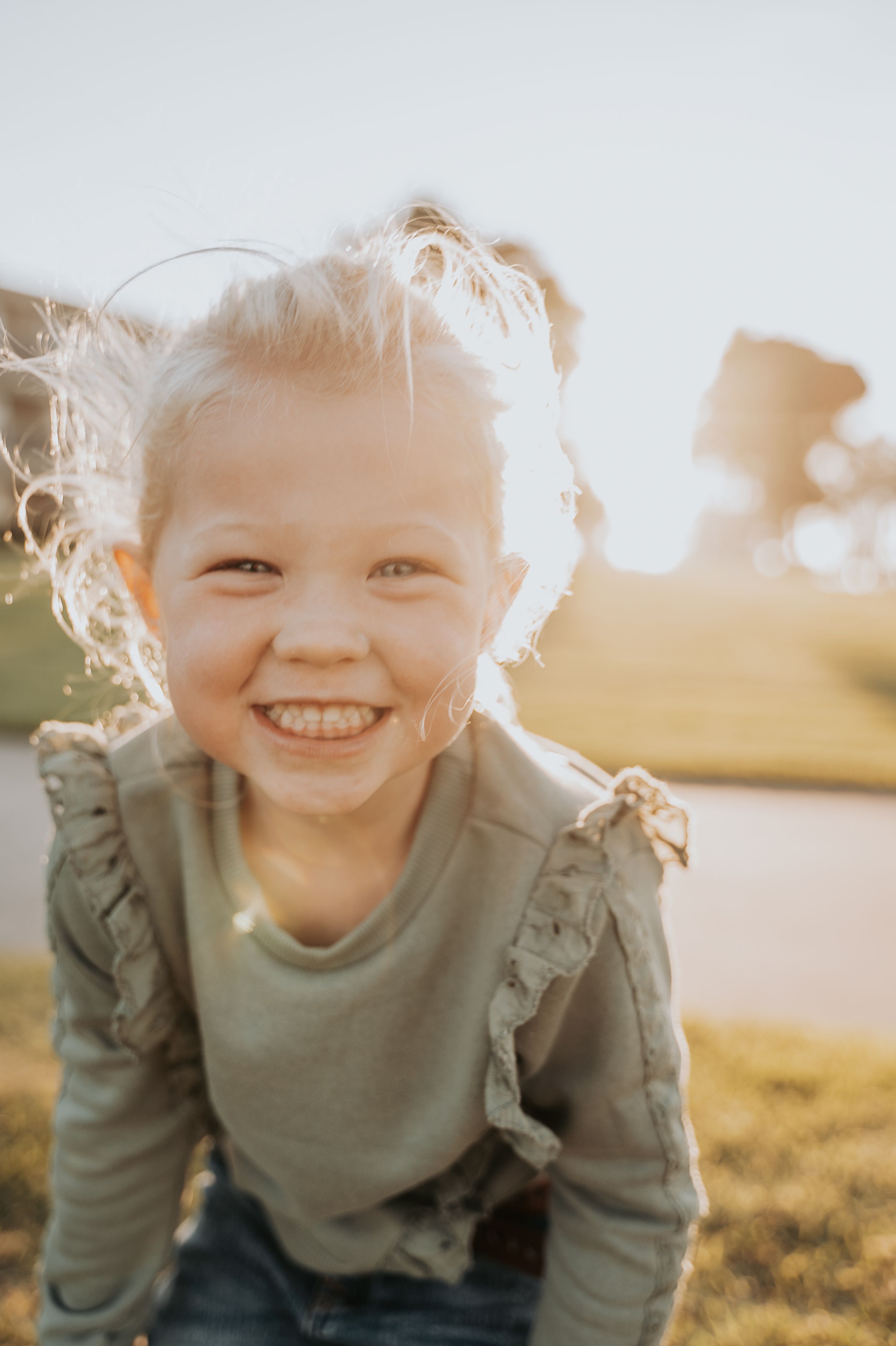 A child smiles brightly at the camera while playing outdoors in the golden sunlight.