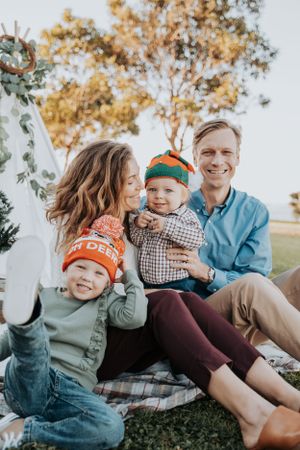 A family sits together outdoors wearing winter hats and casual clothing on a plaid blanket.