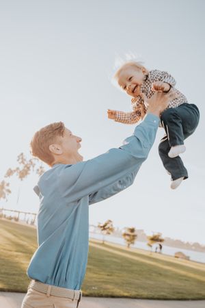 A person lifts a small child high into the air against a bright sky background.