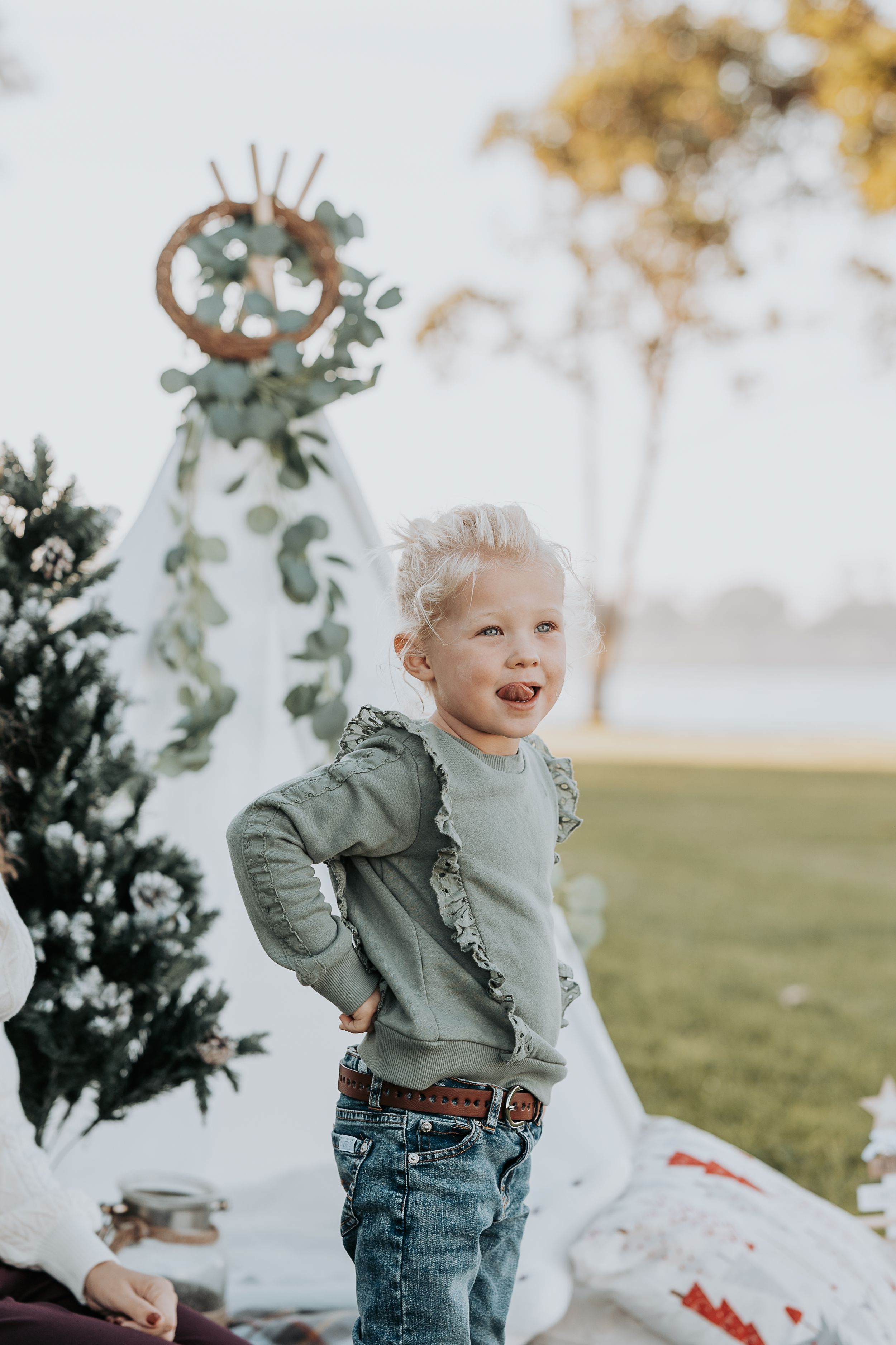 Young child in sage green shirt laughing next to decorated Christmas tree outdoors.