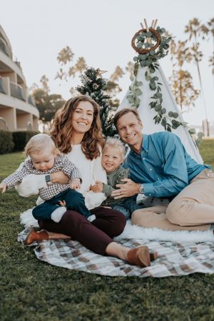 Family sitting on picnic blanket with decorated Christmas tree outdoors.