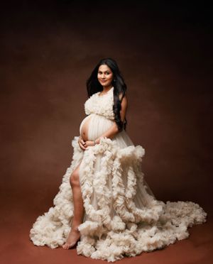 Studio maternity portrait in cream ruffled gown against brown backdrop.