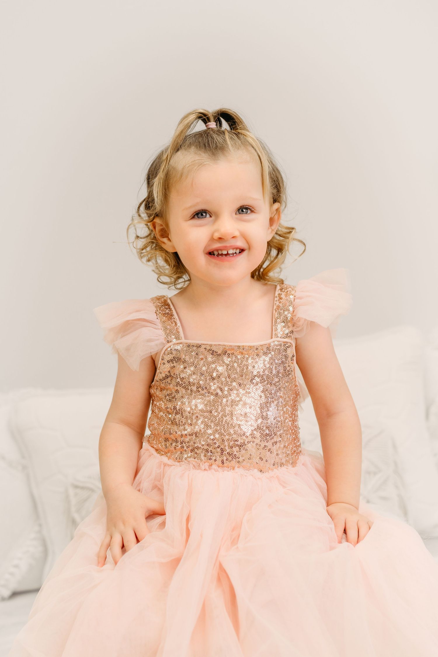 Child in rose gold sequin and tulle dress smiling on white couch.