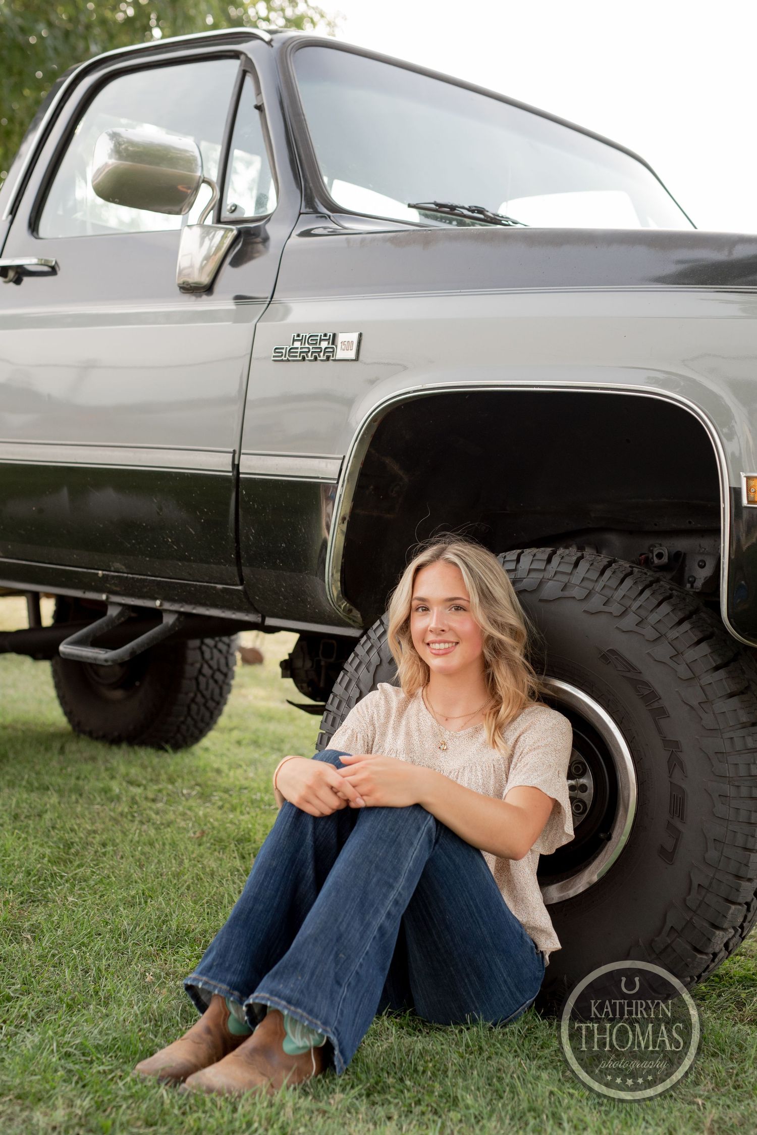 Someone relaxes against the wheel of a lifted GMC pickup truck on grass.