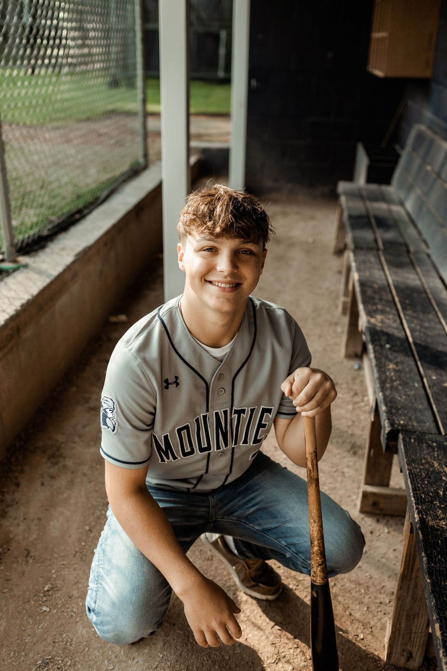 A baseball player wearing a Mount jersey sits in a dugout with a bat.