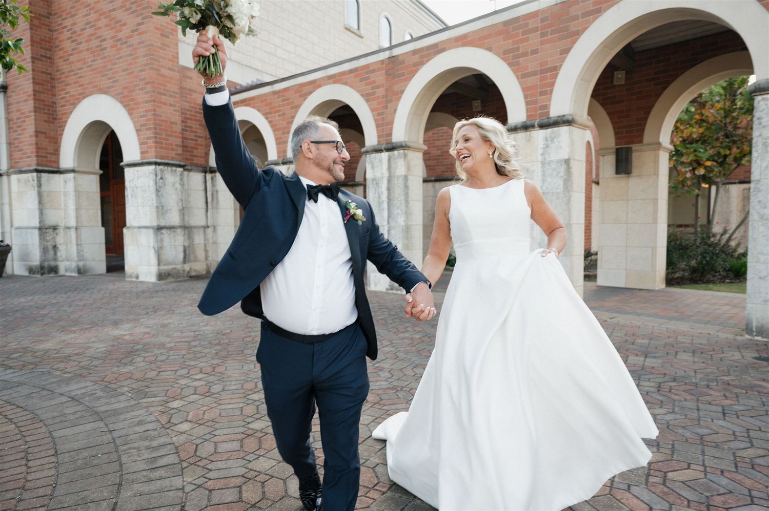 Newlyweds celebrate with raised arms under brick archways in courtyard.