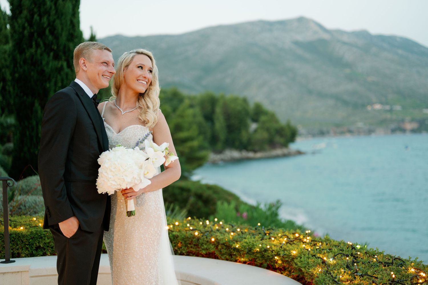 A couple in wedding attire embraces on a scenic overlook with mountains and blue water in the background.