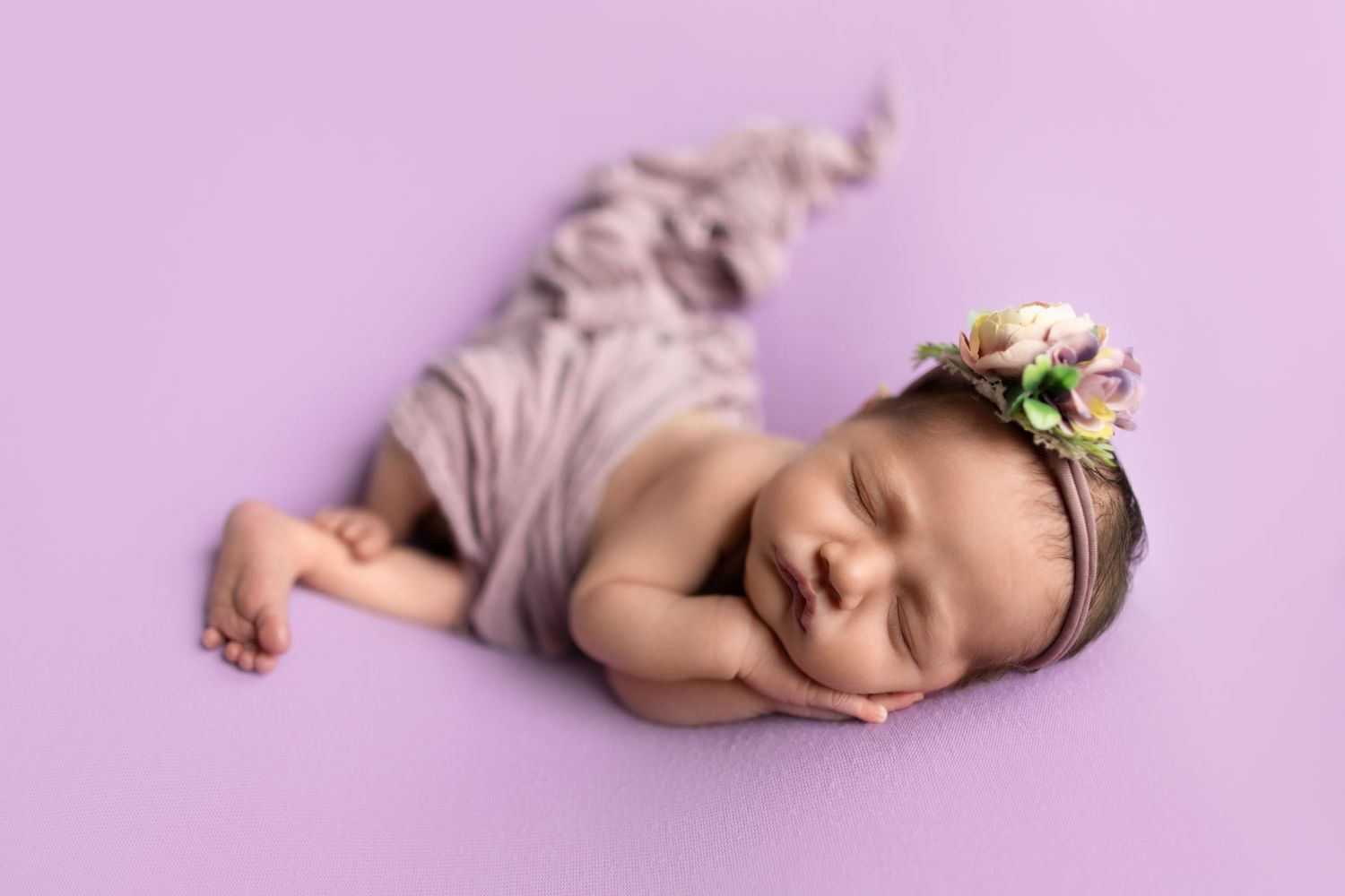 Sleeping baby in floral headband posed on purple background.