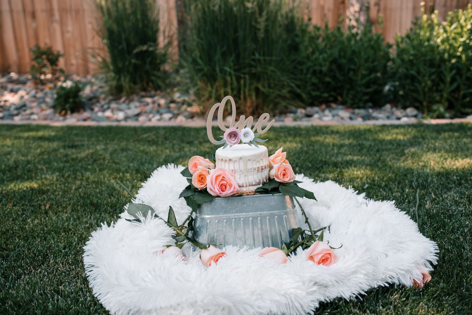 A white cake decorated with pink roses and 'One' text on a fluffy white surface in a garden setting.