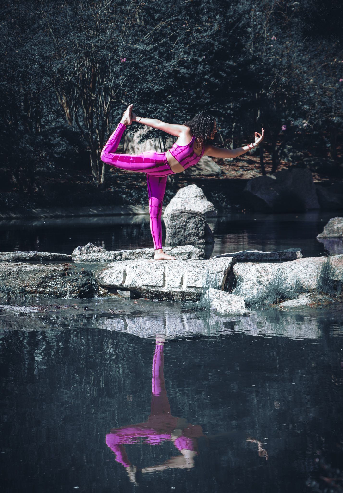 Yoga practitioner in bright pink attire strikes a pose on rocks with reflection in still water.