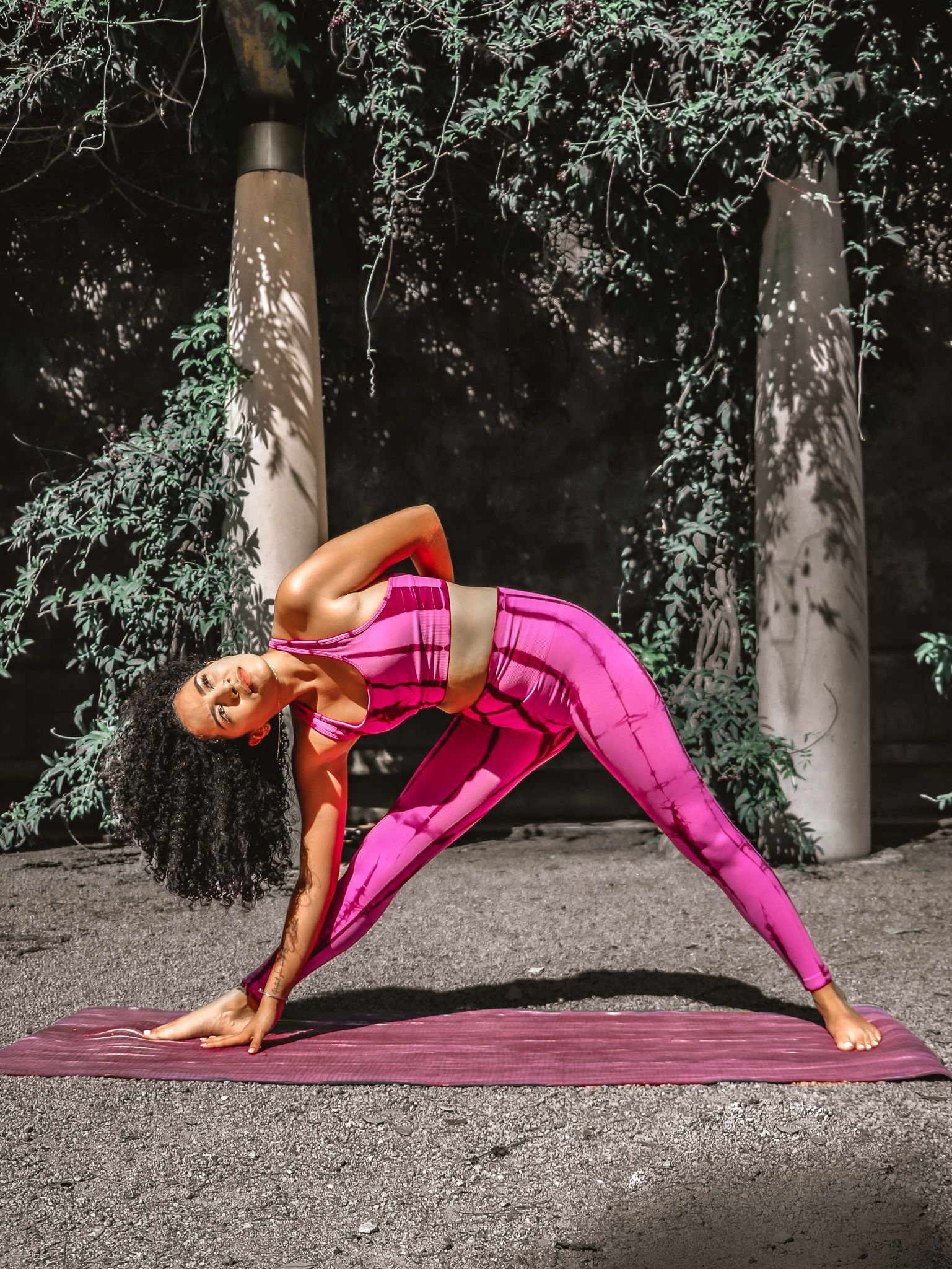 Yoga pose demonstration in bright pink athletic wear against dark foliage background.