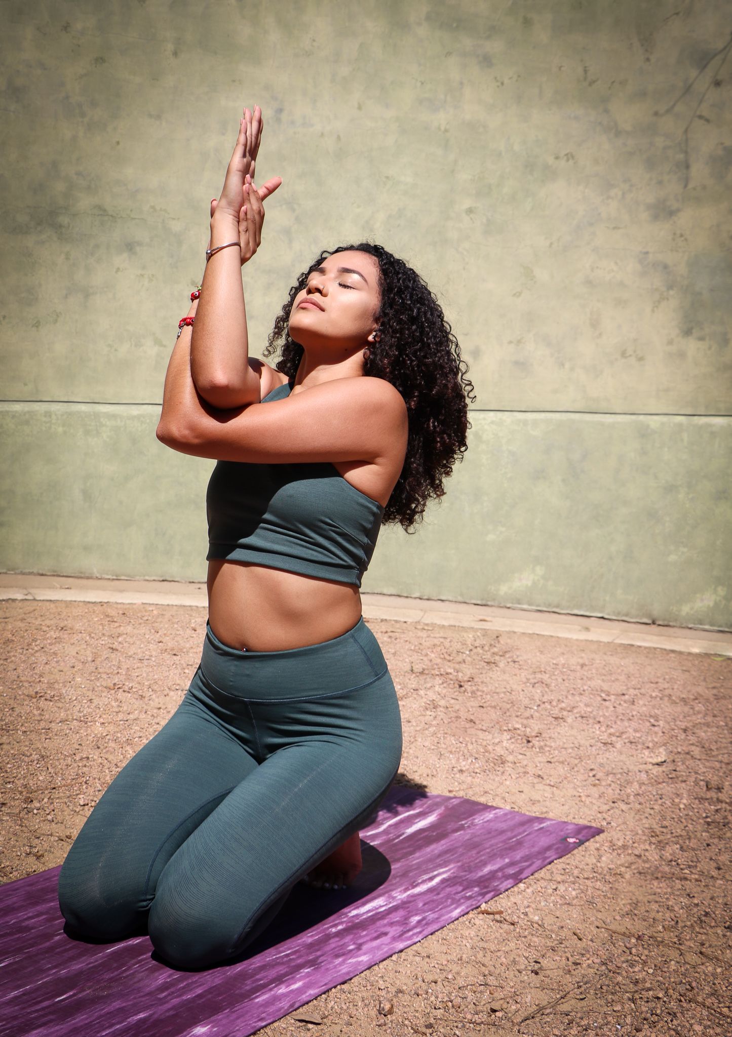 Yoga practitioner in gray athletic wear performs kneeling pose on purple mat outdoors.