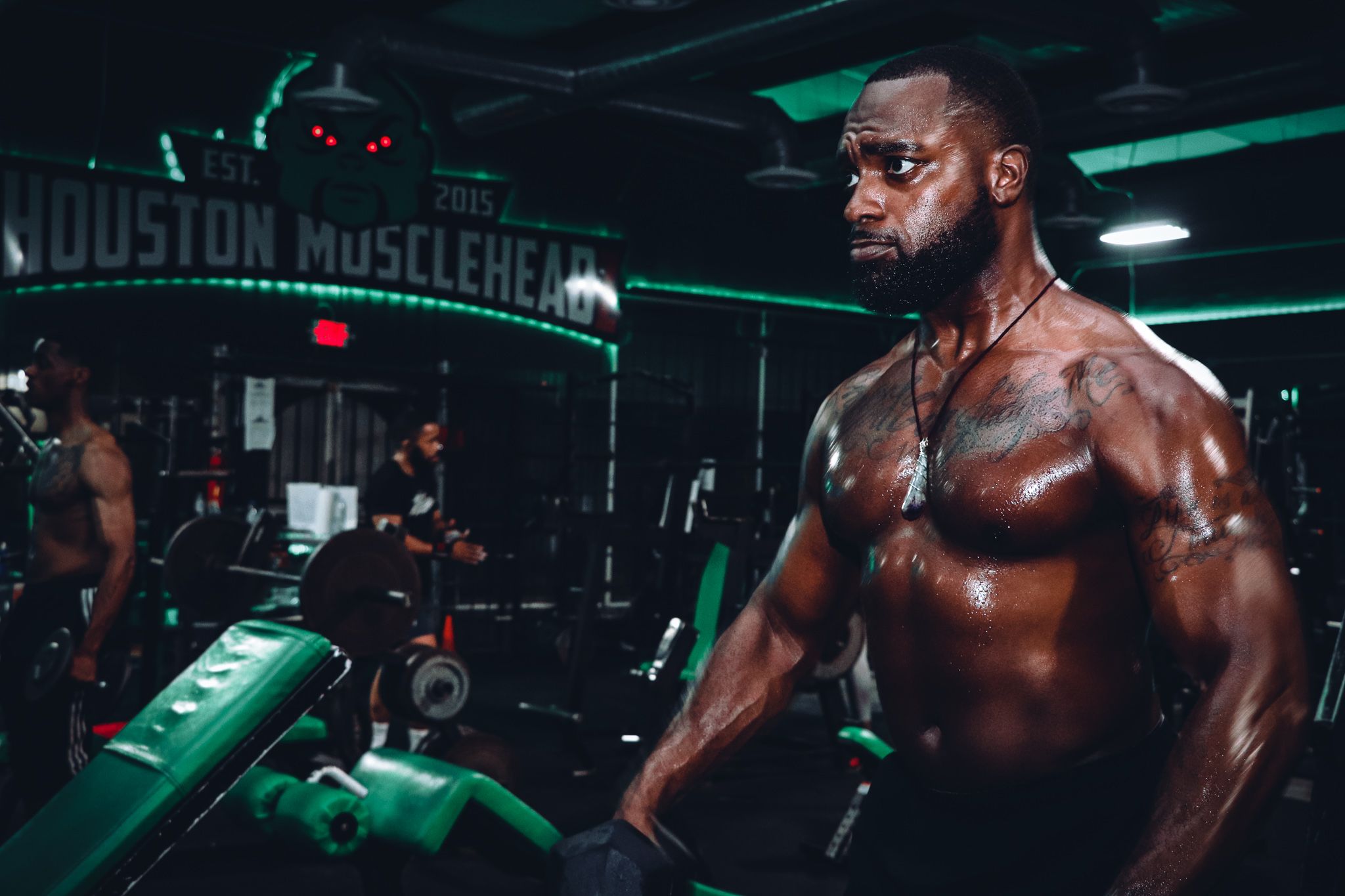 Muscular athlete training at Houston Muscle Heads gym with green lighting and exercise equipment.