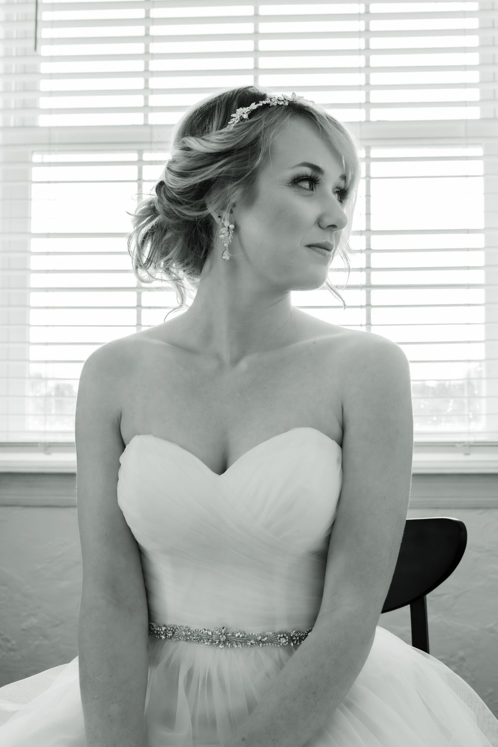 A bride in an elegant white strapless gown with crystal belt poses by a window.