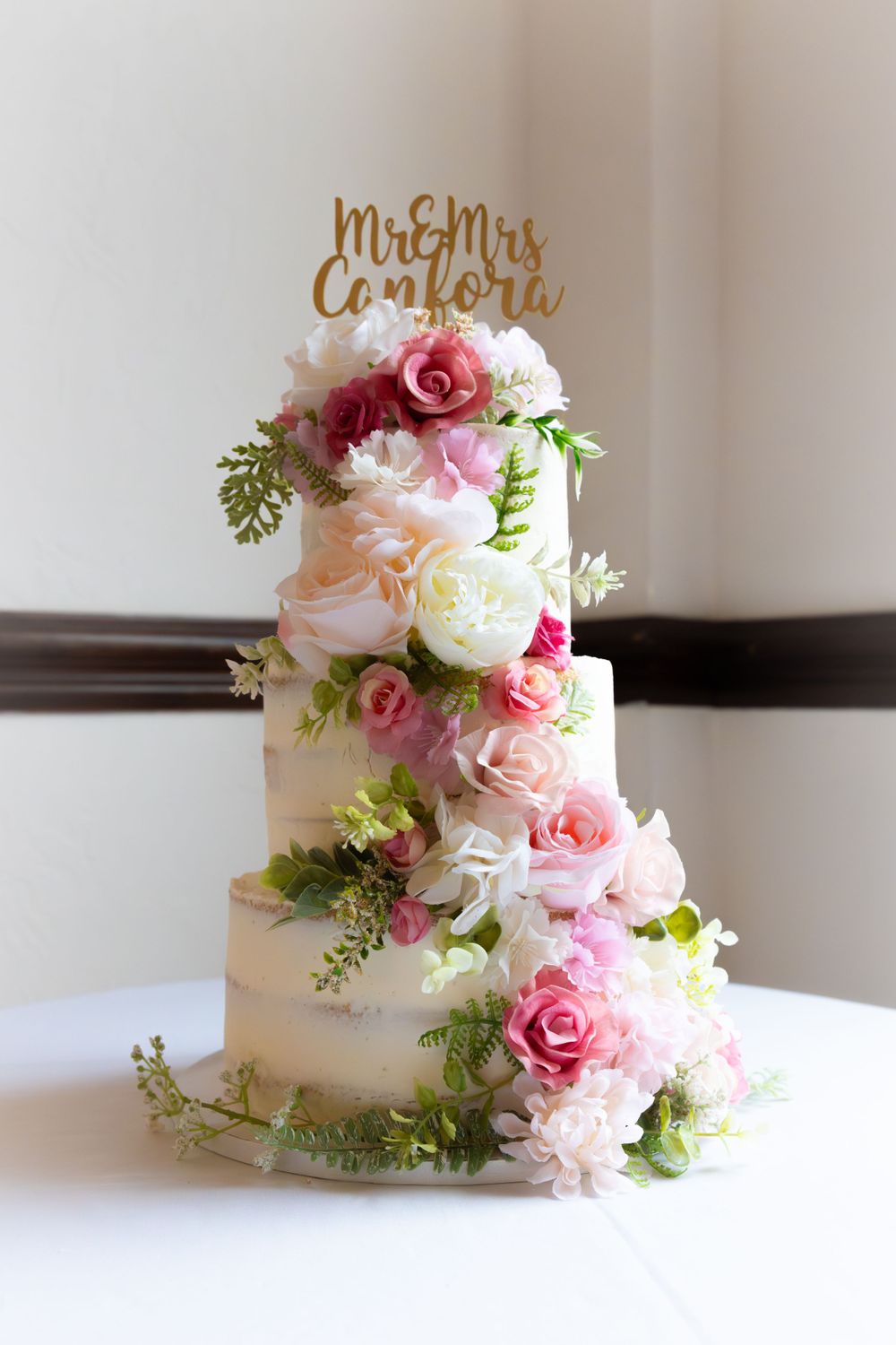 Elegant three-tier white wedding cake decorated with pink and white roses and a gold topper.