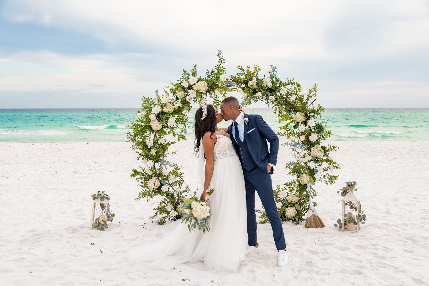 Romantic moment under floral arch on white sand beach with turquoise waters in background.