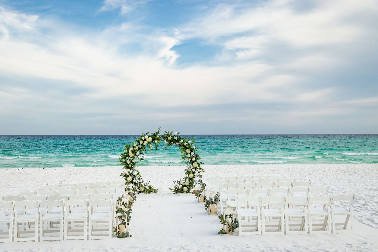 Beach wedding setup with white floral arch and chairs on pristine sand overlooking turquoise ocean.