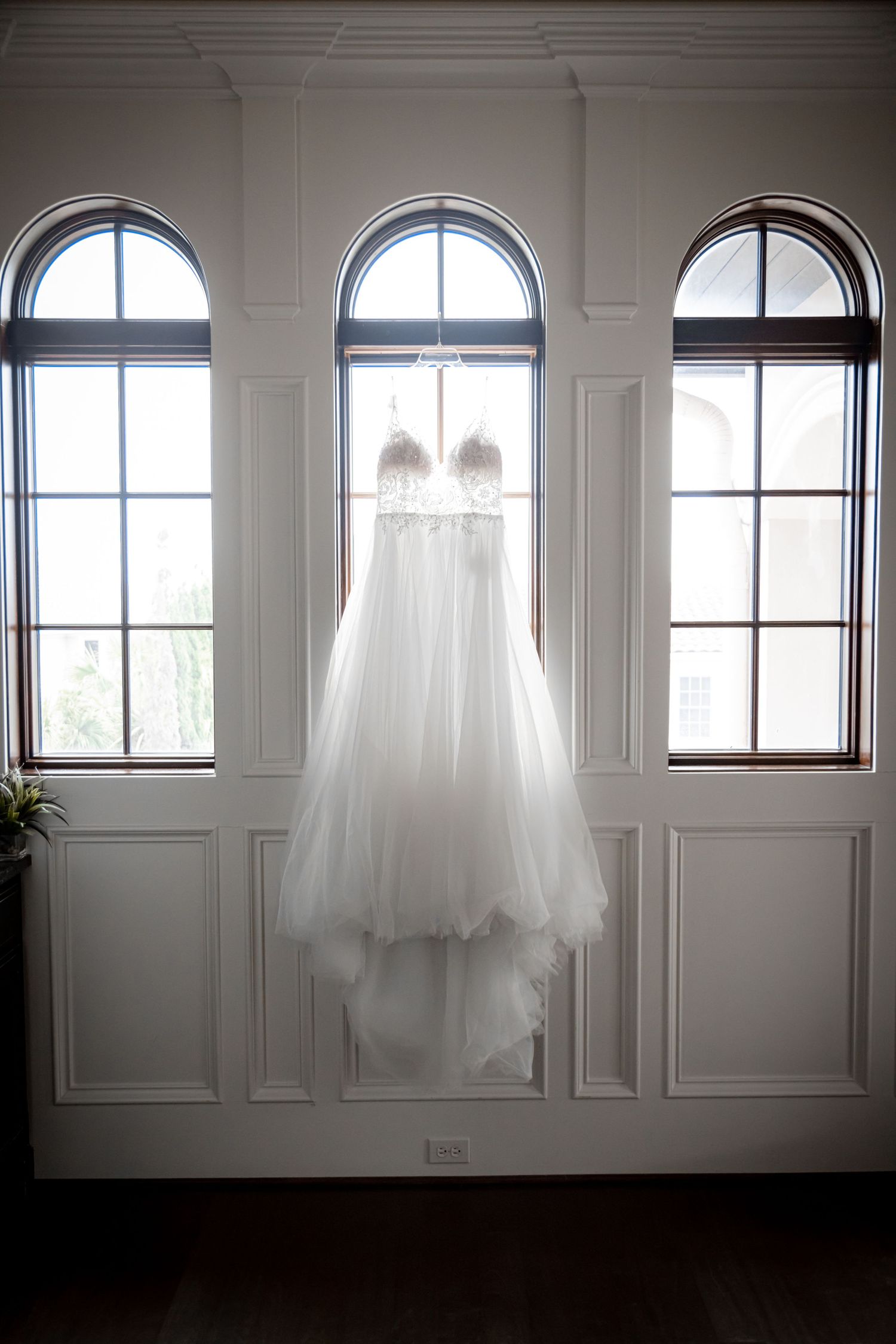 Elegant white wedding dress hangs in front of arched windows creating a dreamy silhouette.