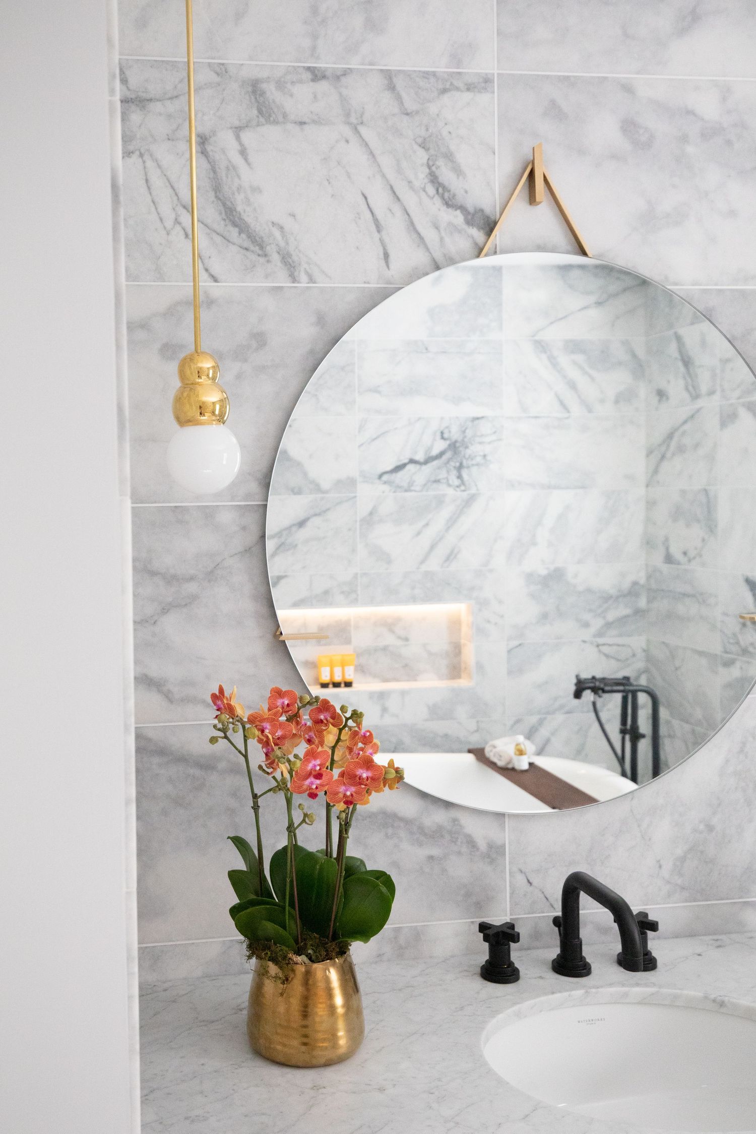 Elegant bathroom with round mirror, marble tiles, and coral orchids in gold vase.