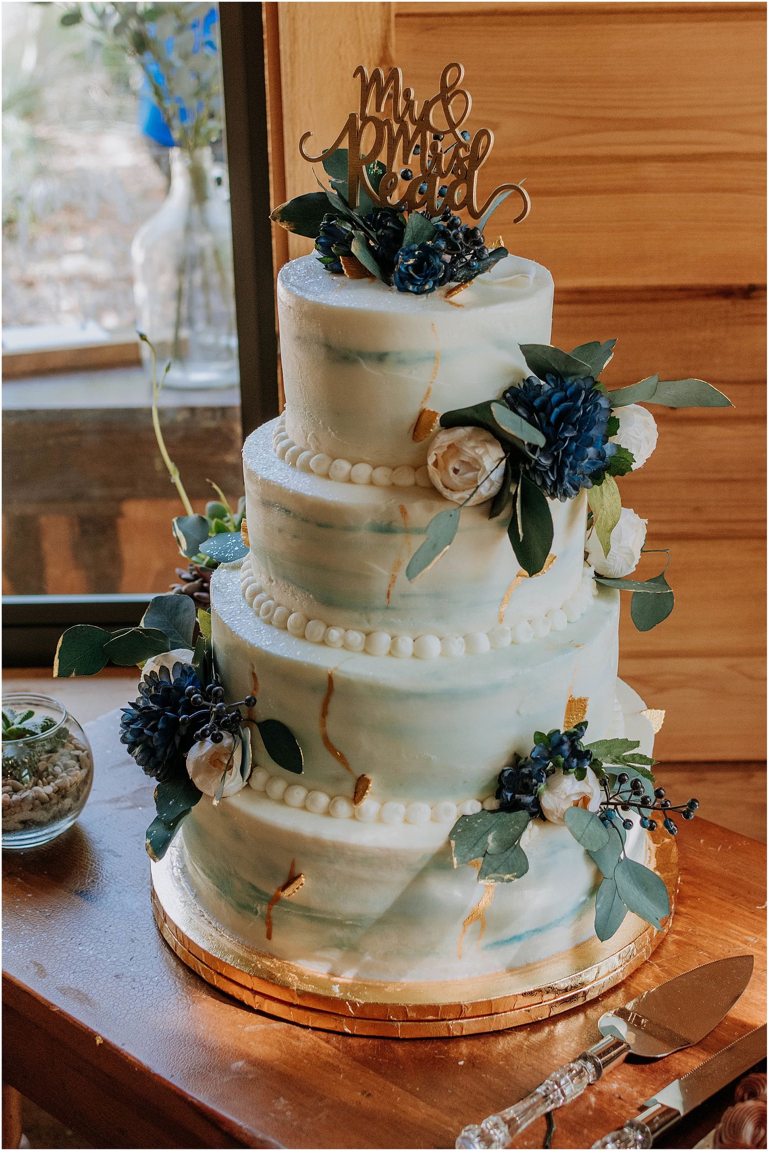 Four tier white marble effect wedding cake decorated with blue flowers and gold cake topper on wooden table.