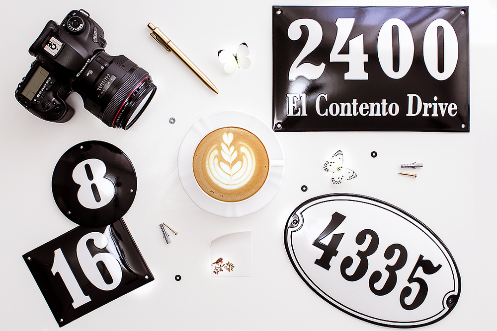 Creative arrangement of camera equipment, coffee cup and vintage-style address numbers on white background.