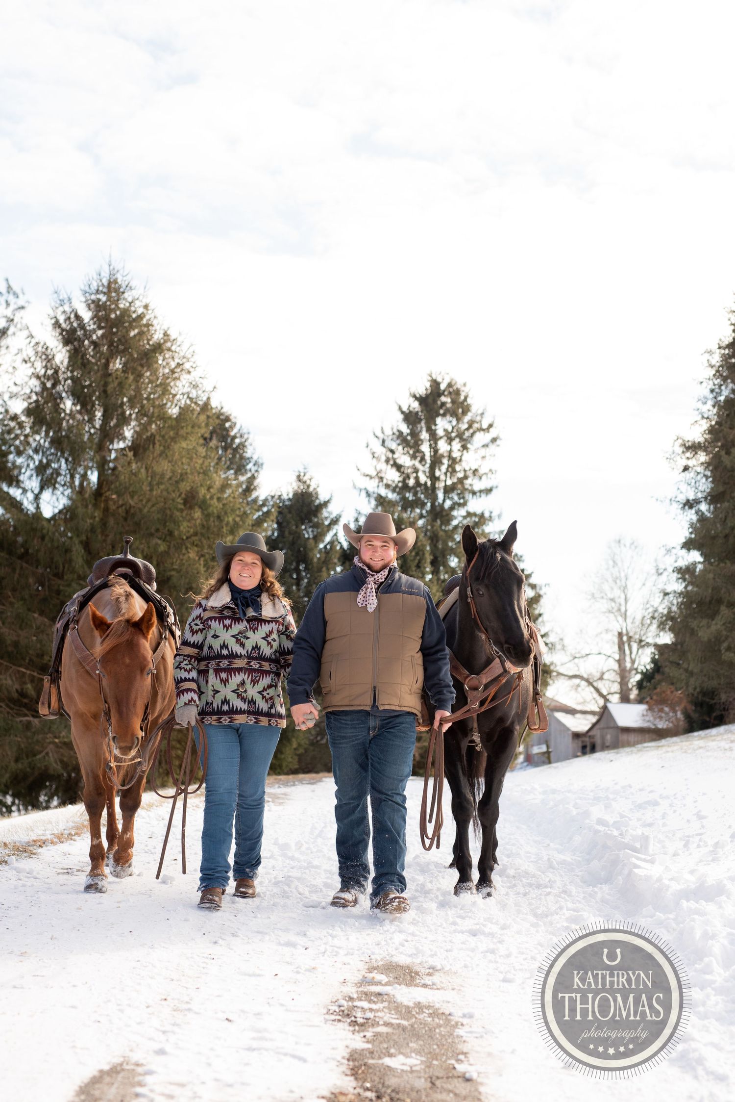 Two horses and their handlers walk together down a snowy winter trail bordered by evergreen trees.