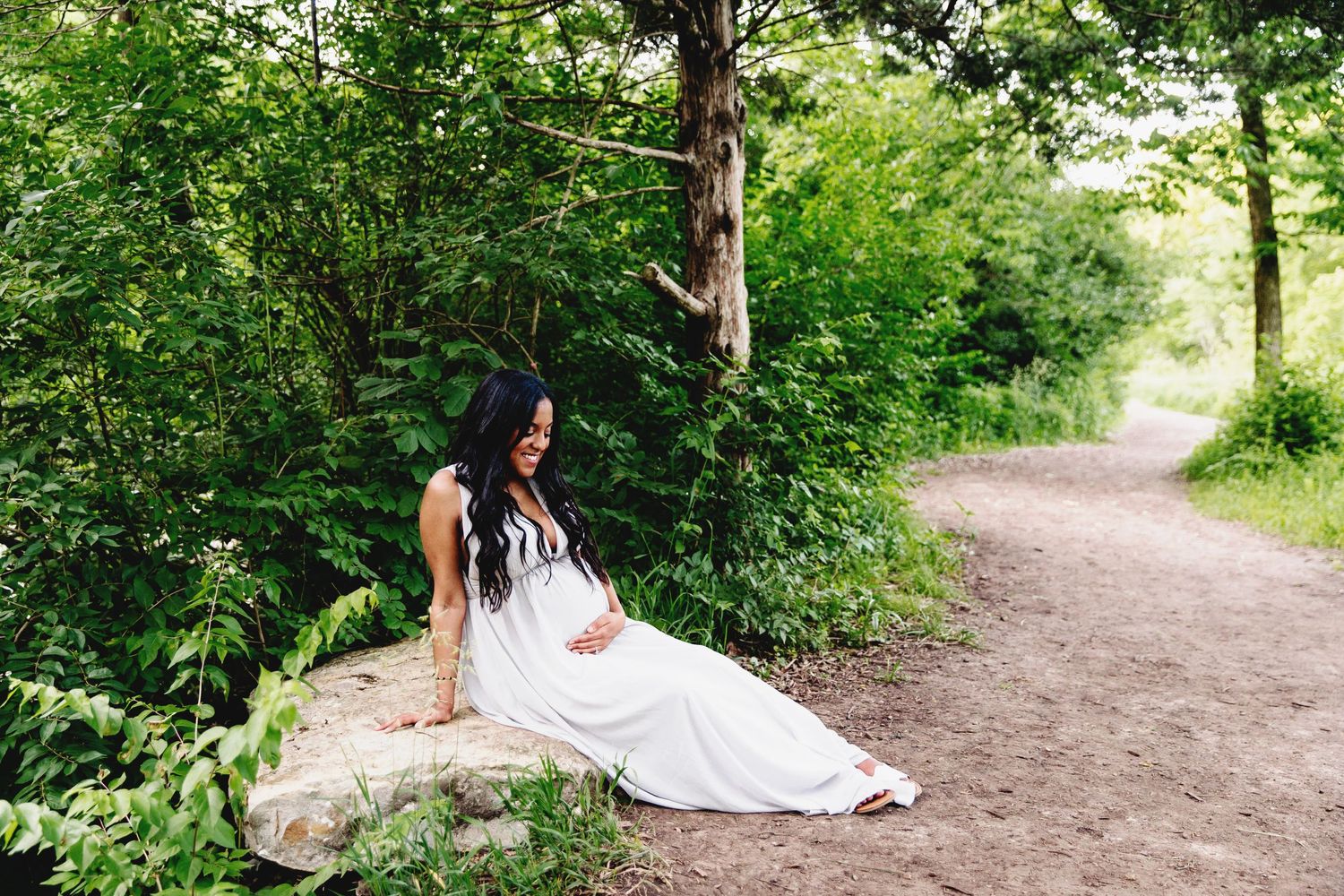 woman sitting on large rock slab near walking path holding her pregnant belly