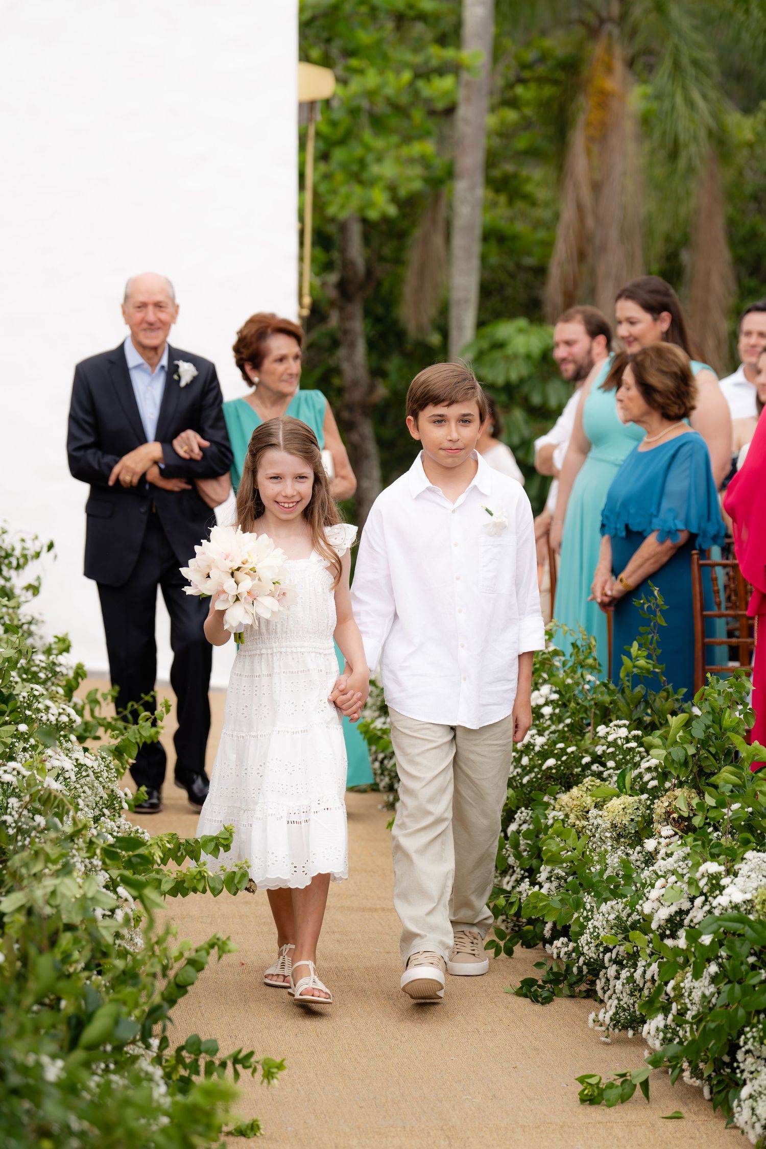 Children walk down flower-lined aisle at outdoor wedding ceremony.