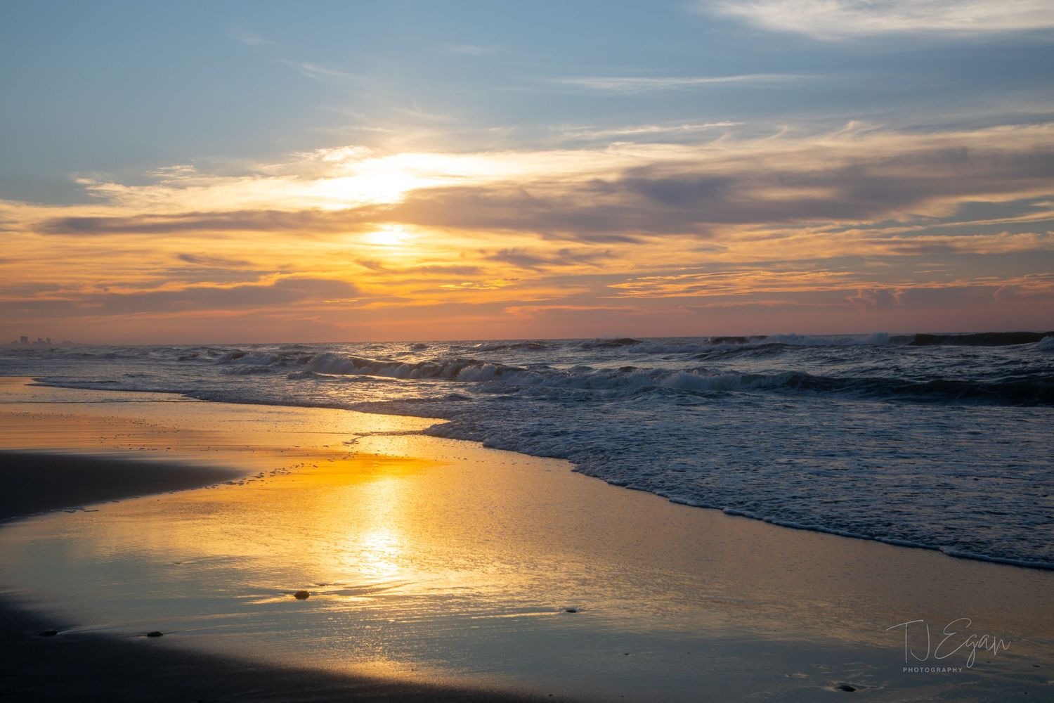 Golden sunset reflects on wet beach sand with rolling waves in background.