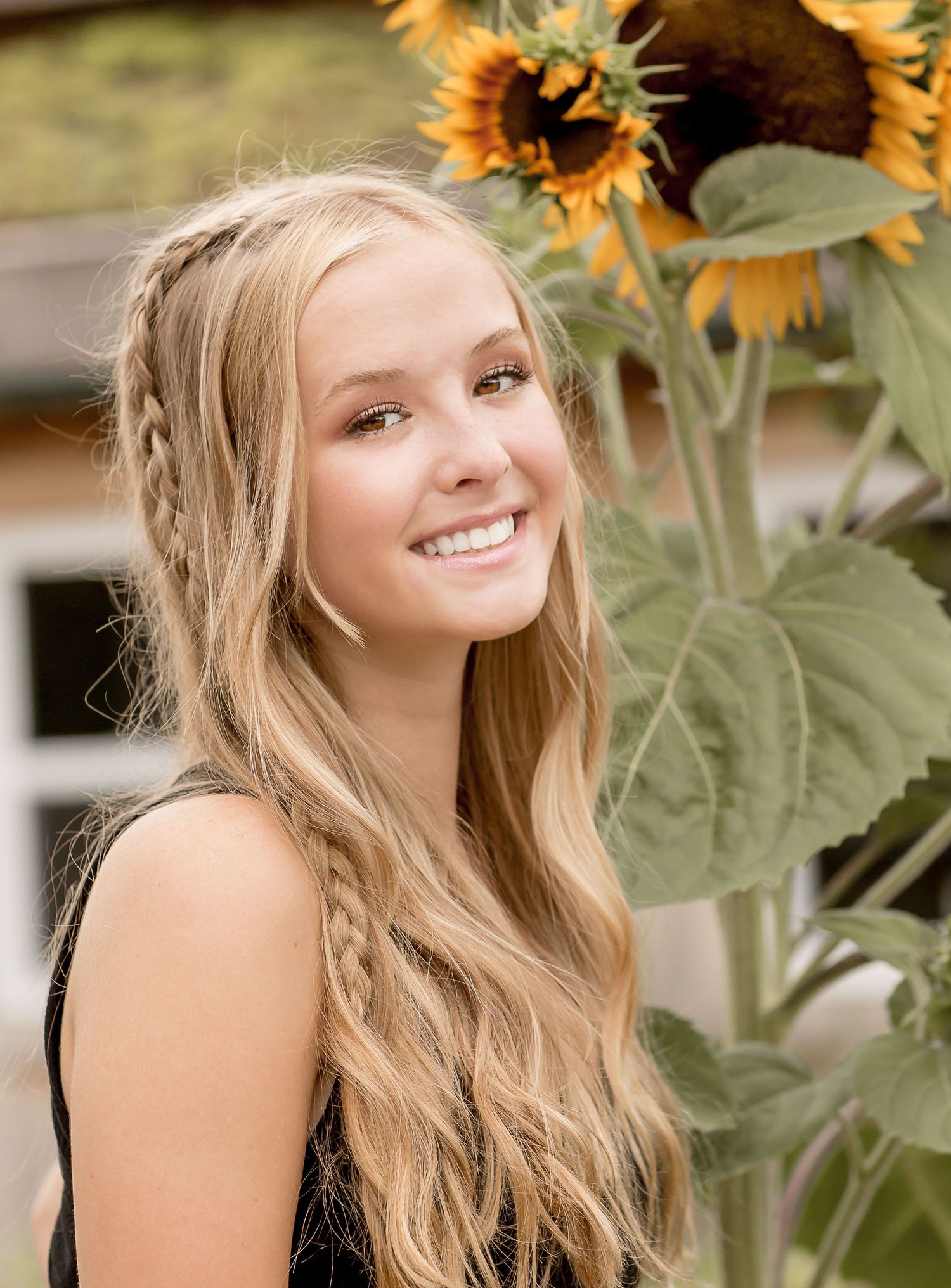 Teenage girl with long blonde braided hair smiling beside tall sunflowers outdoors.