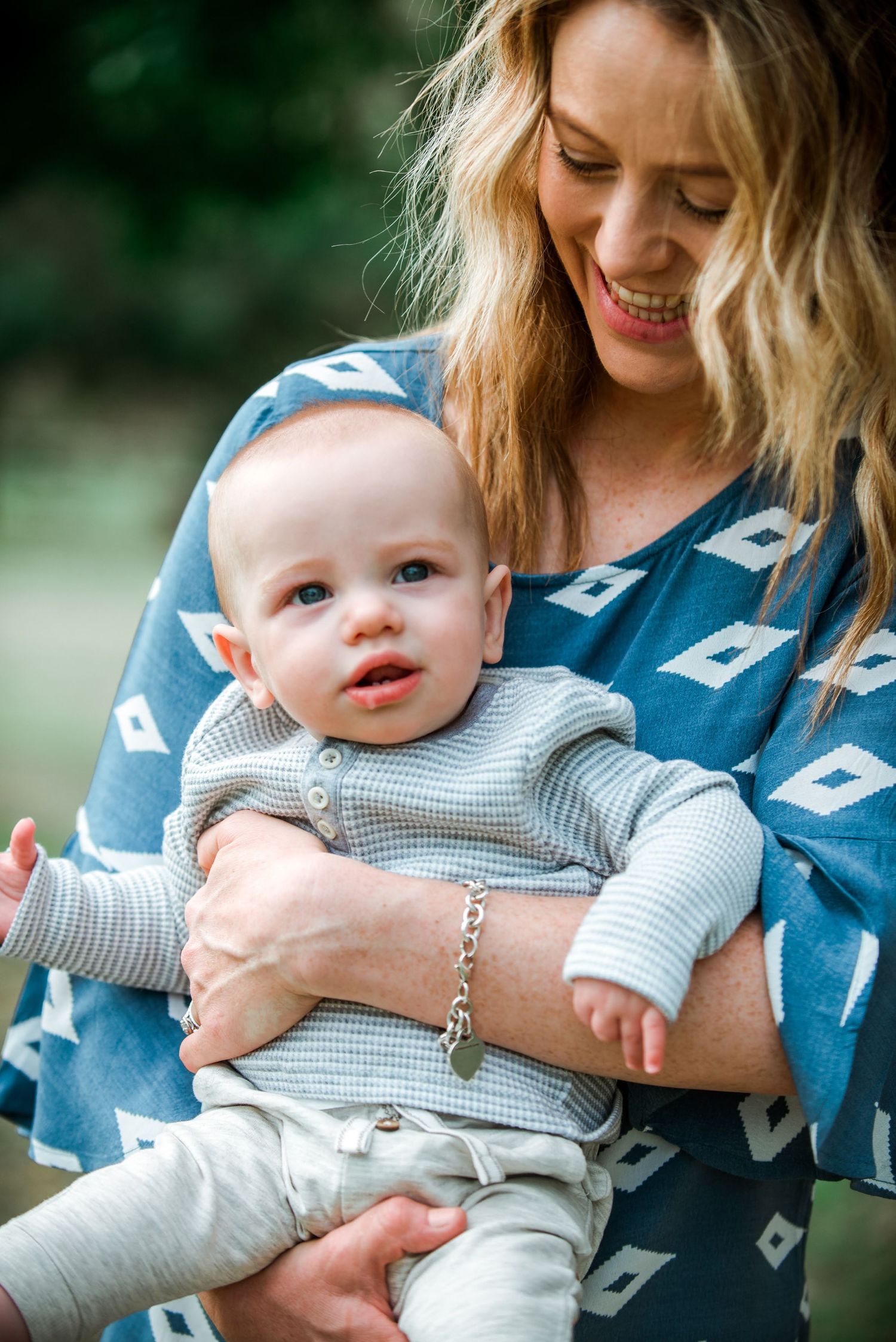 Happy mother in blue patterned dress holding infant in grey outfit during outdoor photoshoot.