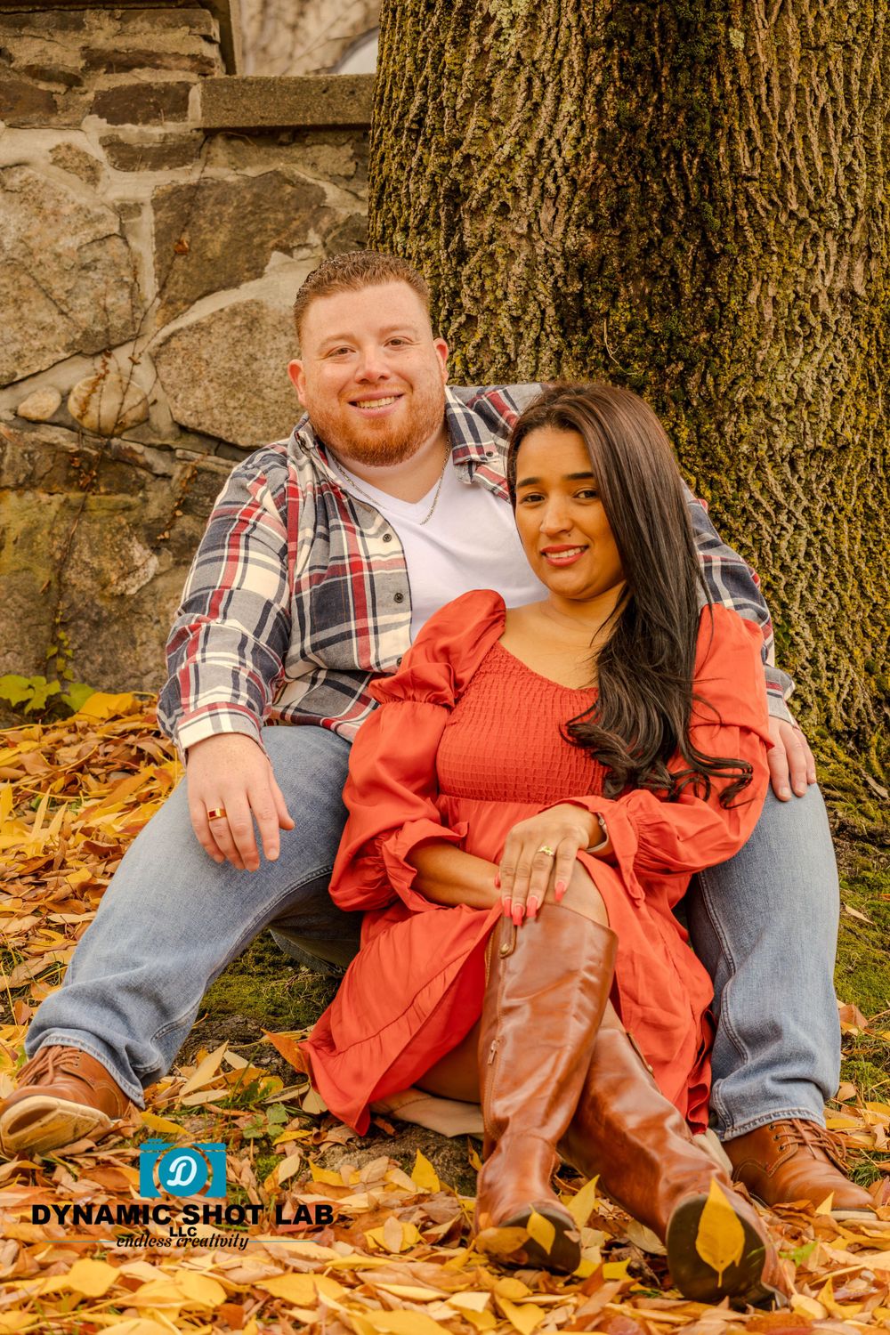 A couple sits together on fallen autumn leaves near a large tree and stone wall.