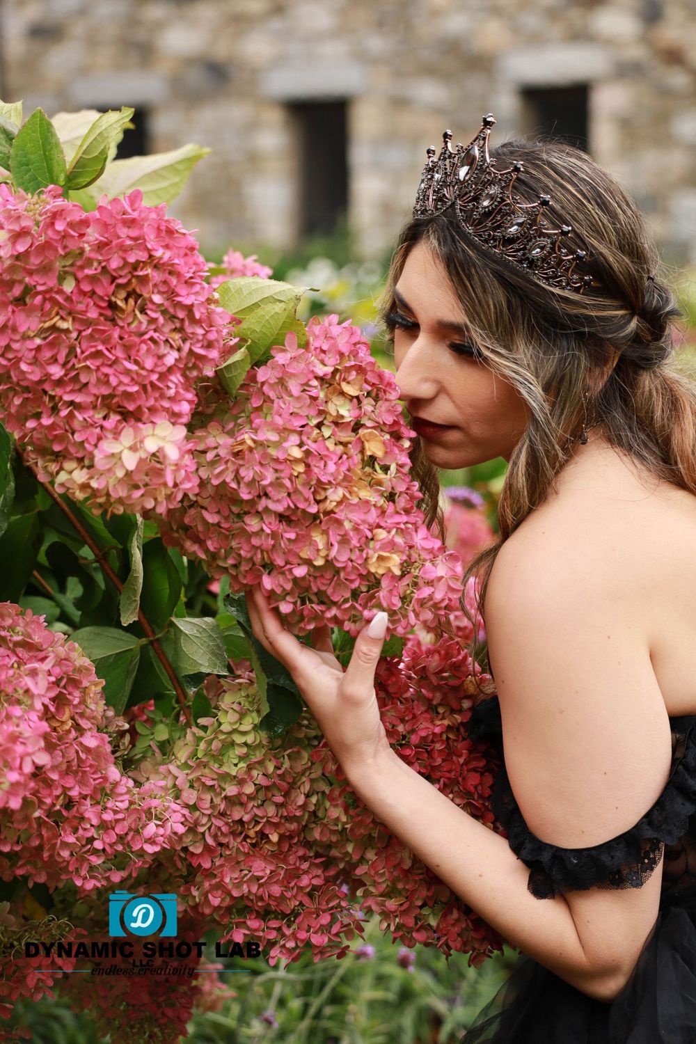 A person in a black dress poses with pink hydrangea flowers against a stone wall.