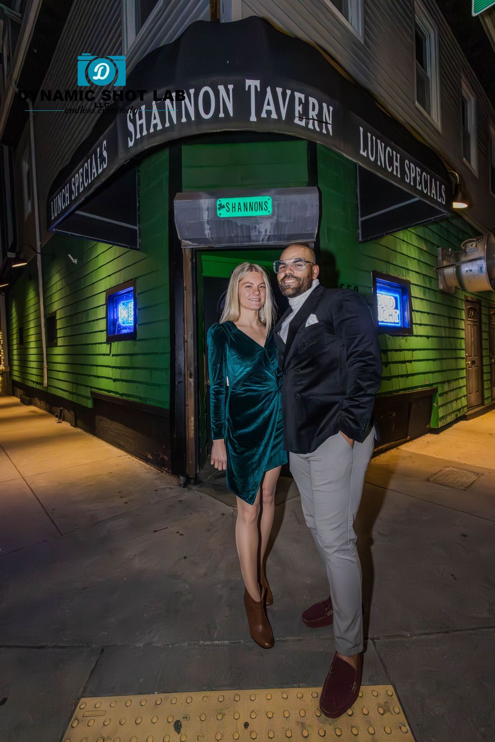 A couple poses playfully outside Shannon Tavern at night with green neon lighting.