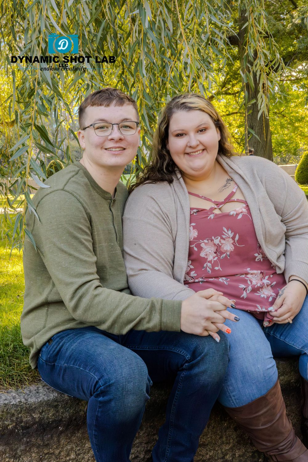 A couple sits together on a stone wall surrounded by autumn foliage wearing casual green and burgundy attire.