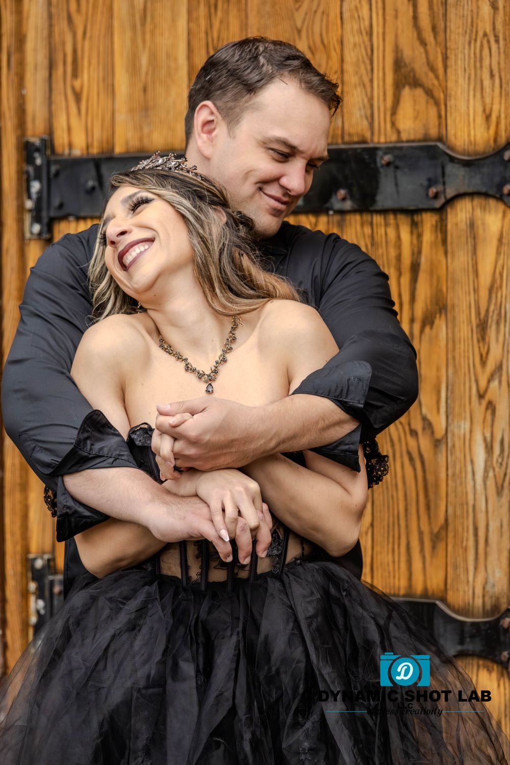 A romantic couple embraces against a wooden wall, wearing black formal attire and sharing a joyful moment together.