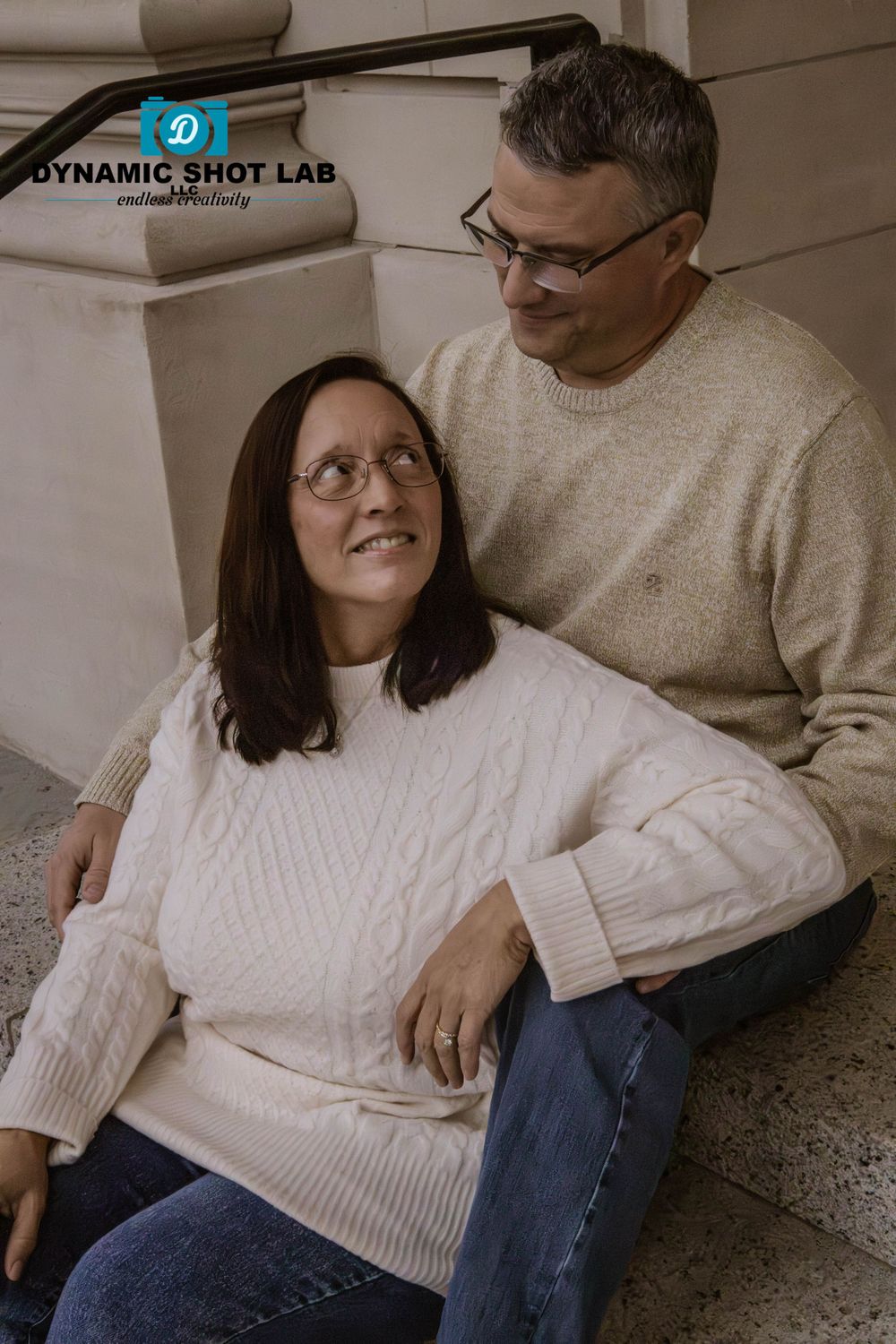 A couple relaxes on a couch wearing cozy white sweaters and jeans in an intimate indoor setting.