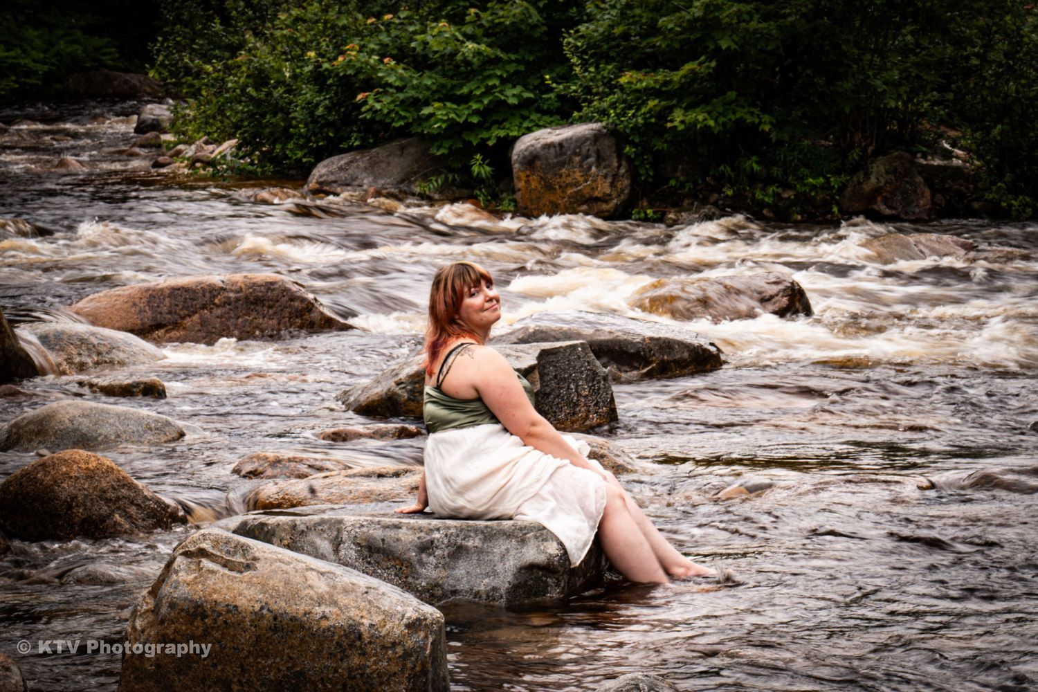 Woman in white dress sitting peacefully on rocks beside a rushing river surrounded by lush greenery.